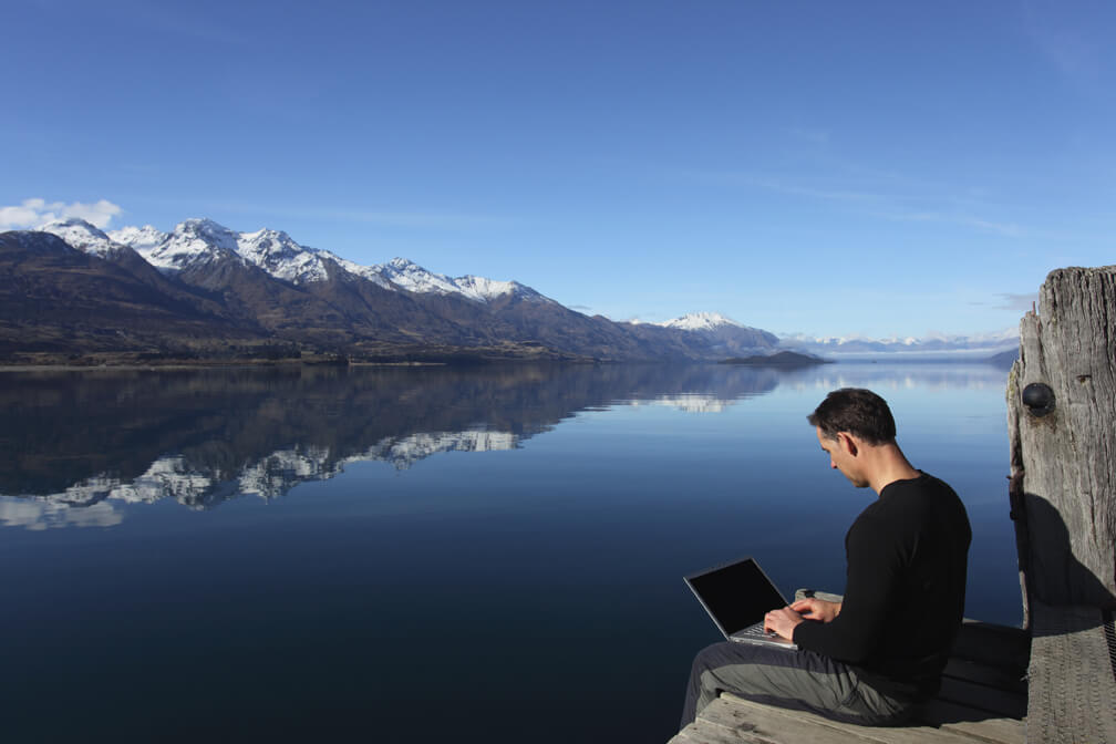 Um homem sentado a beira de um lago com várias montanhas com picos congelados a frente