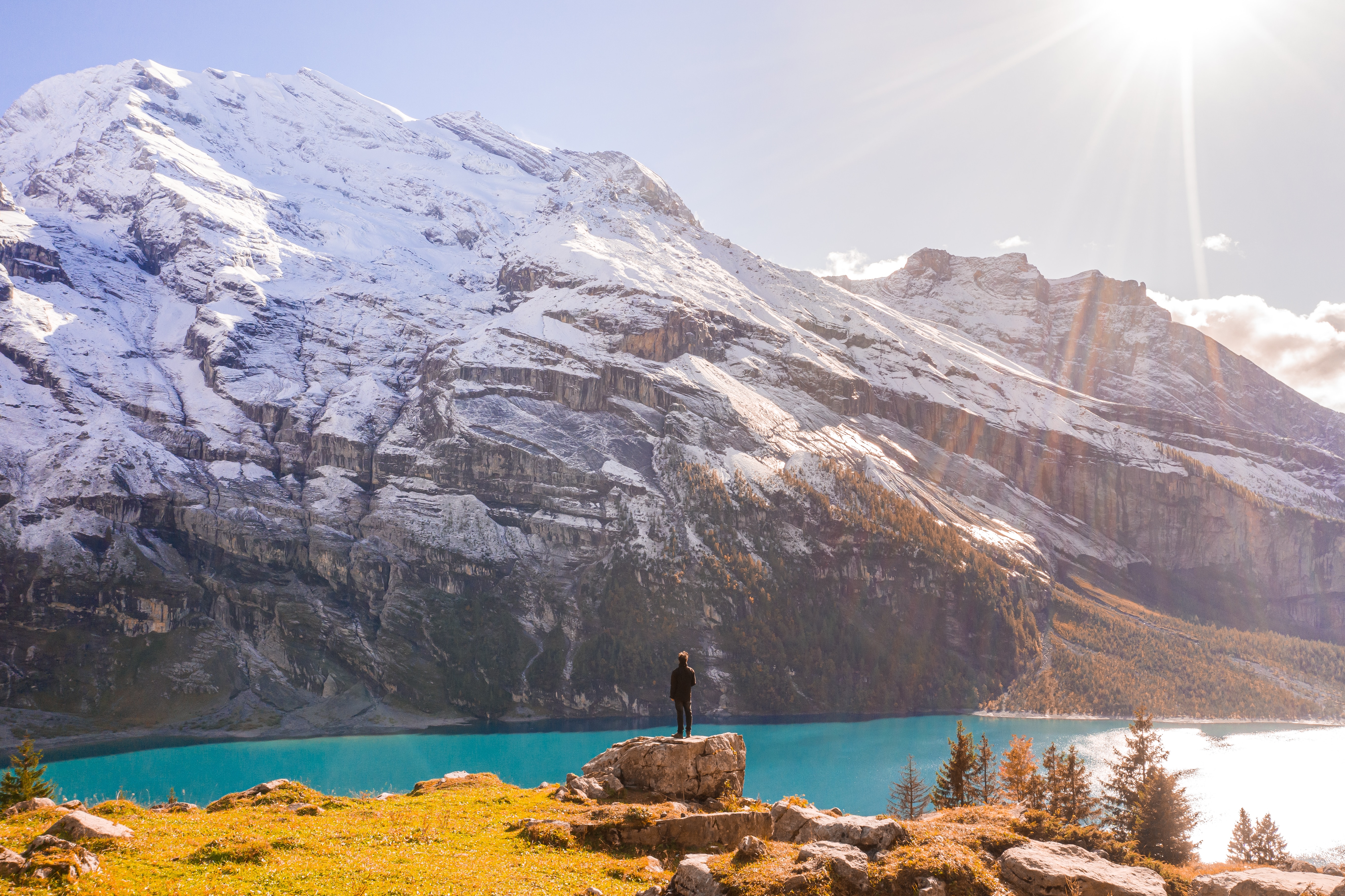 Foto de montanhas nevadas ao fundo, um pequeno trecho de lago super azul e uma pessoa de costas em pé numa pedra.